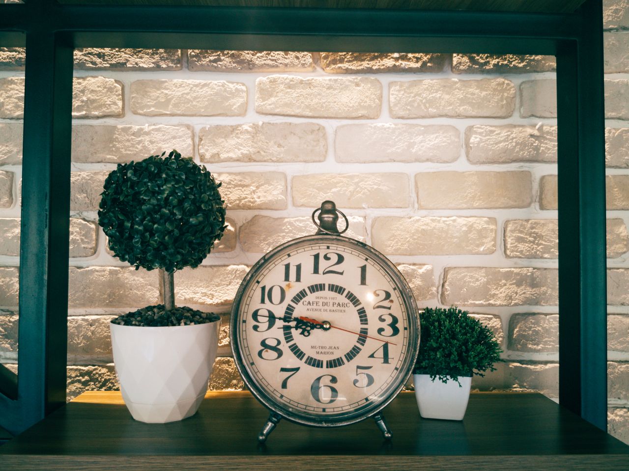 Rustic analog clock with potted topiary plants on wooden shelf against brick wall.